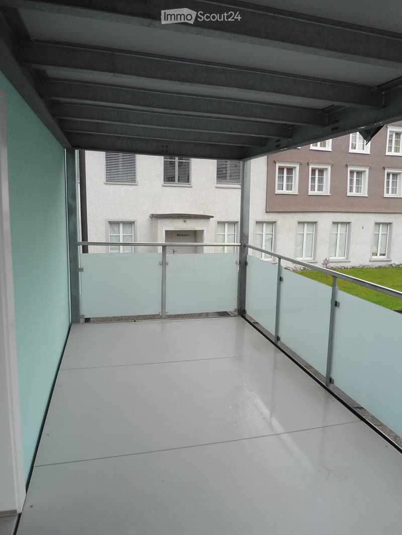 Modern balcony with white tiled floor, glass railings, and a view of neighboring buildings and greenery.