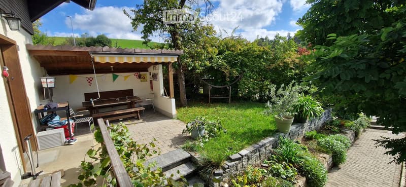 Garden with a covered seating area, picnic tables, chairs, and various plants and trees.