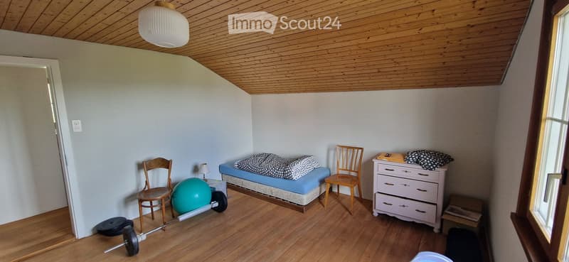 A small, furnished room with wooden flooring and a slanted wooden ceiling. Contains a bed, two chairs, a drawer unit, a fitness ball, and a barbell.