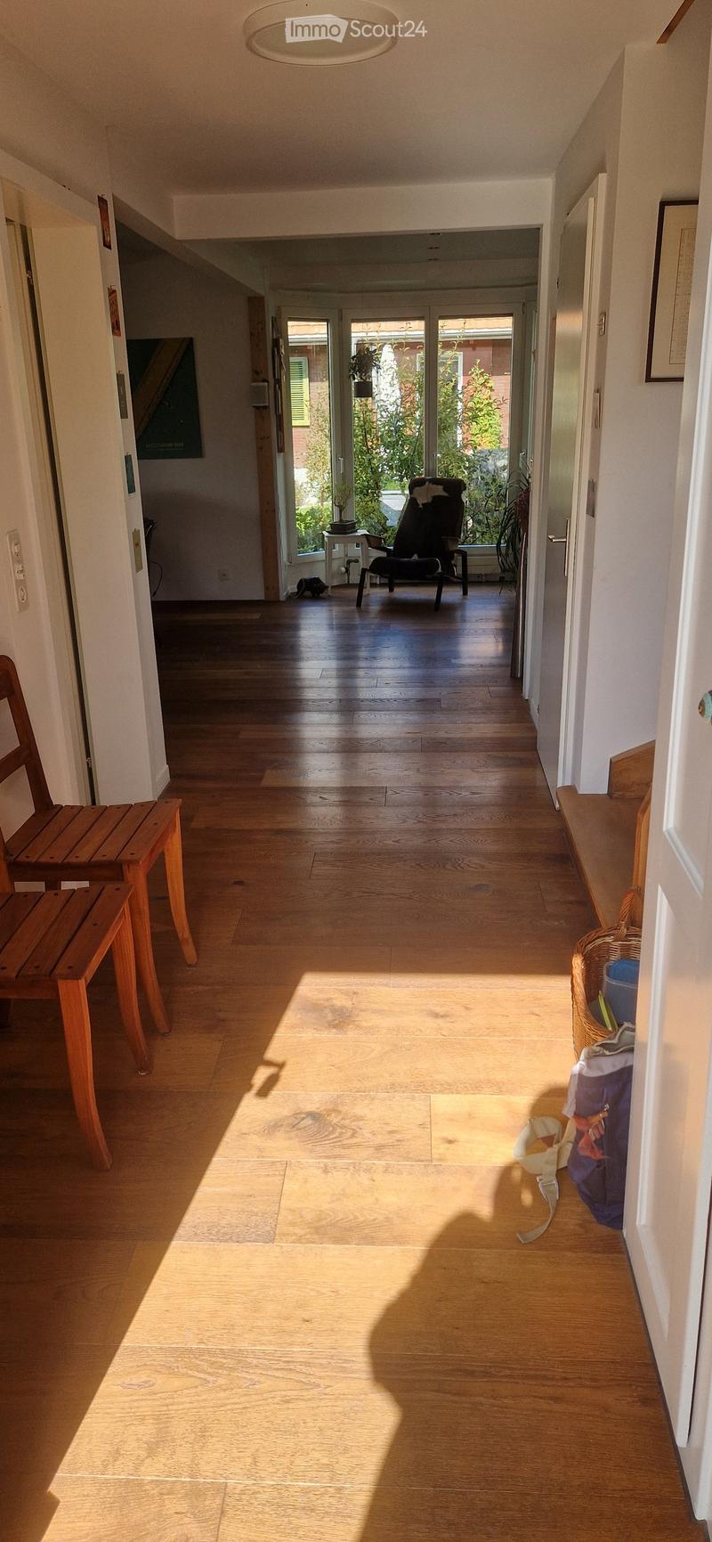 Bright hallway with wooden flooring, white walls, and natural light streaming in from the end. Features a bench on the left, a small table and chair in the adjacent room, and a wicker basket on the right.