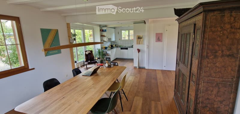 Bright, open-plan living and dining area with wooden flooring, large windows, a dining table with chairs, a laptop, and a kitchen area with white cabinets and a dishwasher.