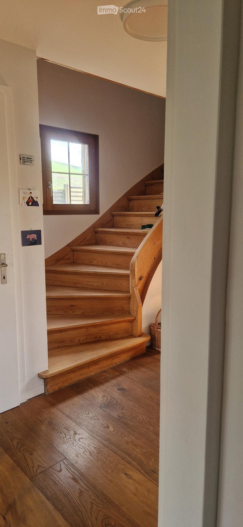 Wooden staircase with a handrail, light-colored wooden steps, and a small window at the top, leading to an upper level. The floor is made of polished wood, and the walls are painted in a light neutral color.