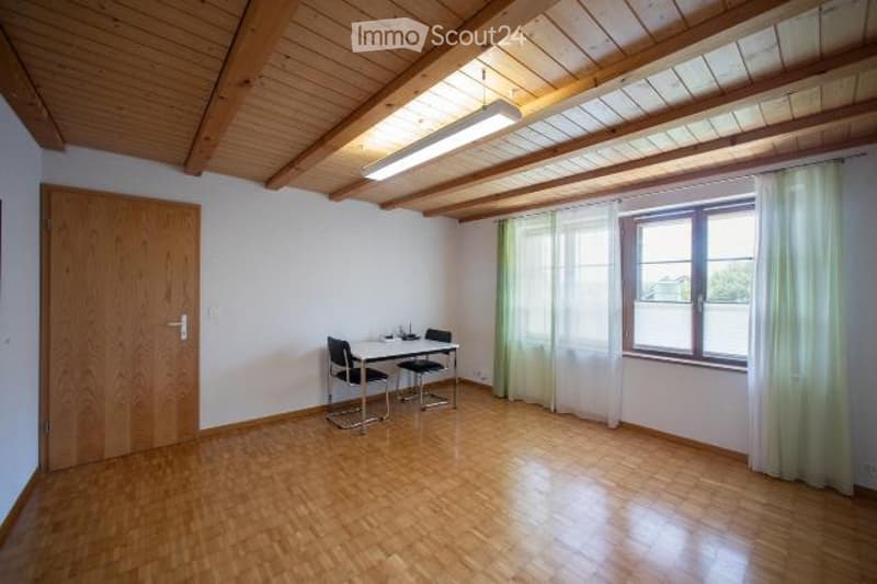 Bright room with wooden floors and exposed wooden beams on the ceiling. Features a small table with two chairs, a large window with white curtains, and a wooden door.