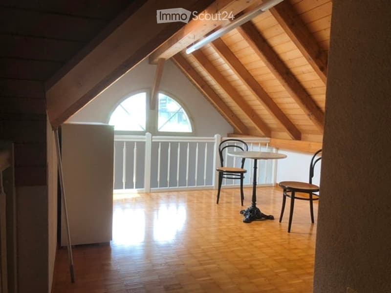 Attic room with wooden beams, slanted ceiling, and exposed wooden floor. Features a small round table with two chairs, a refrigerator, and a white railing along the edge.