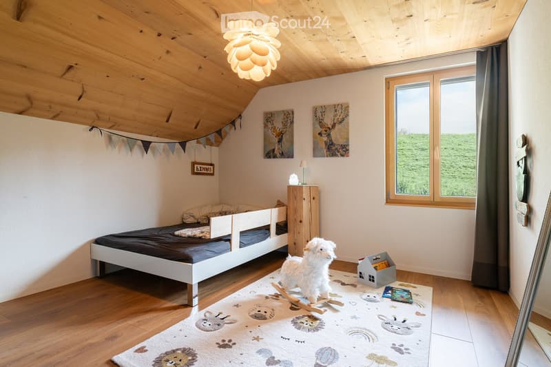 Cozy children's bedroom with a single bed, wardrobe, rocking horse, and colorful rug featuring animal prints. The room has wooden floors, exposed wooden beams on the ceiling, and large windows with curtains.