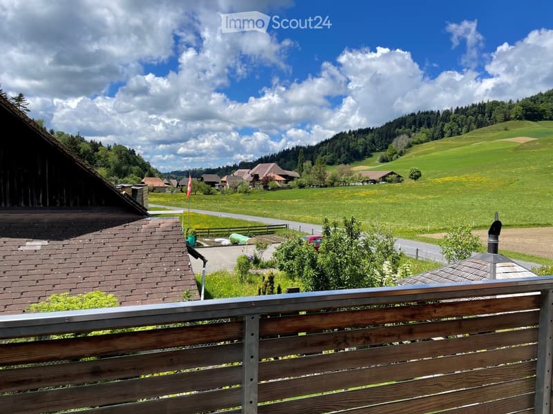 View from a balcony overlooking a rural landscape with green fields, scattered houses, and a road. The foreground shows a wooden railing and part of a roof, with trees and hills in the background.