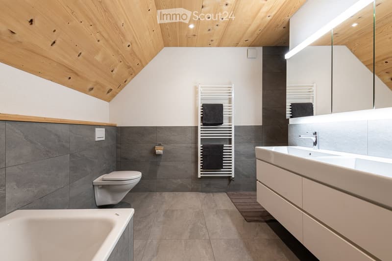 Modern bathroom with grey tiles, white fixtures, a large mirror, and a towel rack with black towels.
