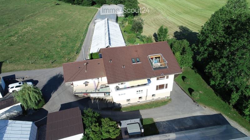 Aerial view of a two-story house with a red-tiled roof, surrounded by green fields and trees. Features include a balcony, a patio area with outdoor furniture, and a driveway with parked cars.