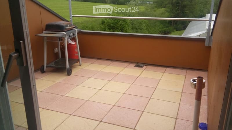Balcony with tiled floor, metal railing, and a small grill cart with a black cover and red container. A cat scratching post is visible in the corner.
