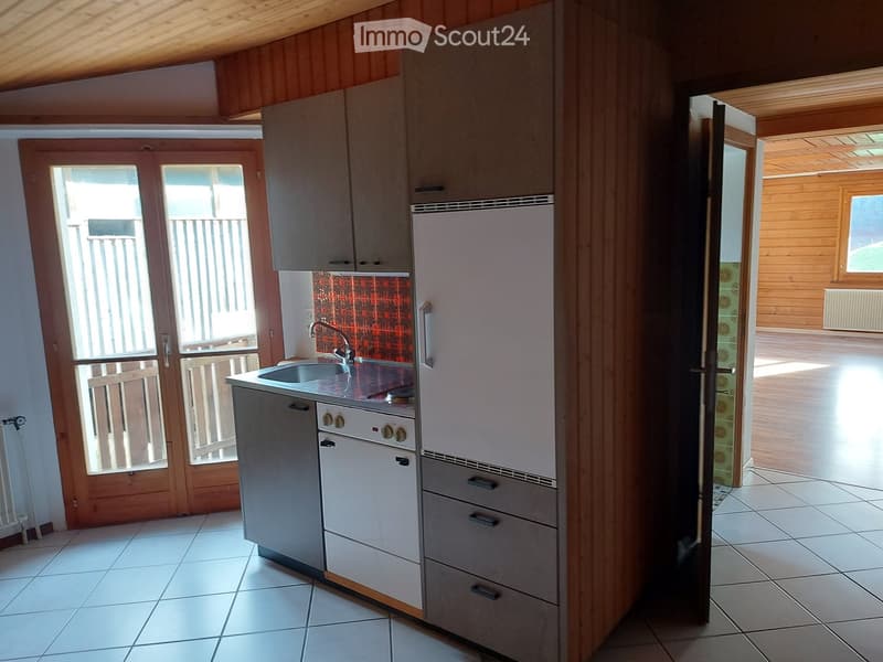 Modern kitchen with white cabinets, stainless steel sink, refrigerator, and oven. Wooden walls and tiled floor, with a view to a wooden-floored living area through an open doorway.