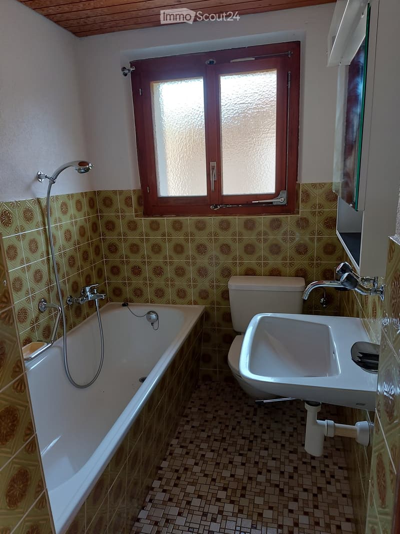 Bathroom with a bathtub, toilet, sink, and mirror. The walls are tiled with a floral pattern, and there is a window with frosted glass panes.