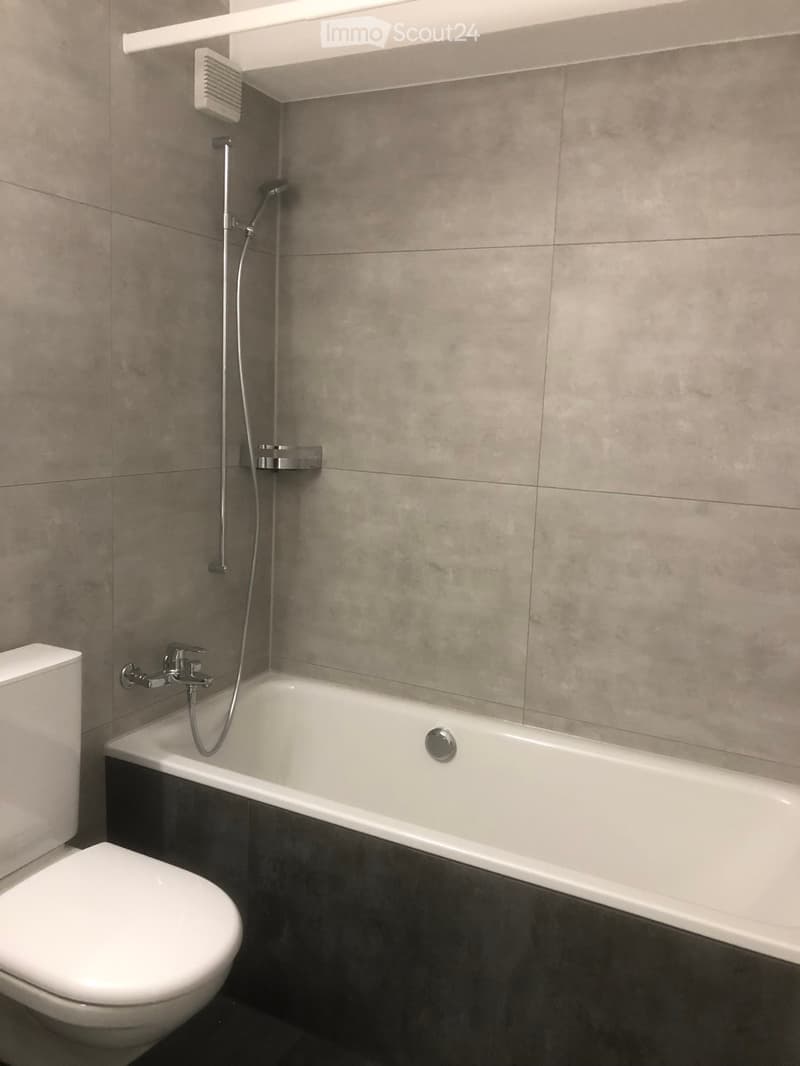 Modern bathroom with a bathtub, toilet, and showerhead, featuring grey tiled walls and a dark grey floor.