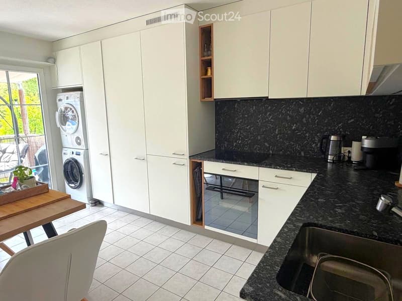 Modern kitchen with white cabinets, black granite countertops, a sink, a washing machine, a small table, and a chair. The kitchen features a built-in oven and a coffee maker on the counter.