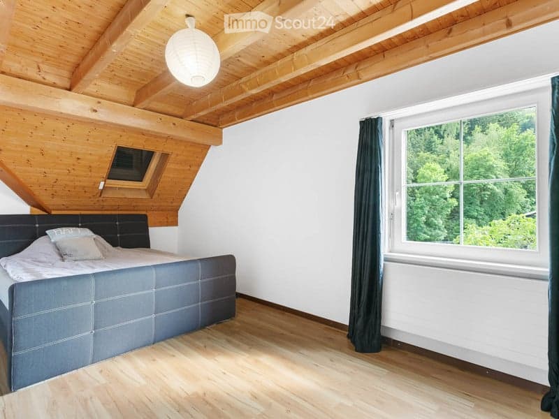 Cozy attic bedroom with wooden slanted ceilings, large window offering a view of greenery, dark grey upholstered bed, and white bedding.