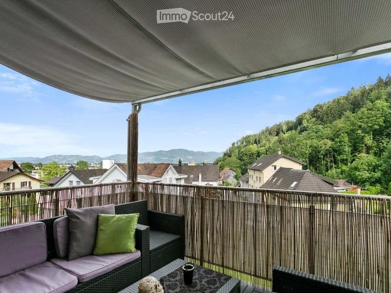 Balcony with a view of the city and surrounding hills, featuring a wicker sofa, cushions, a small table, and a cup.