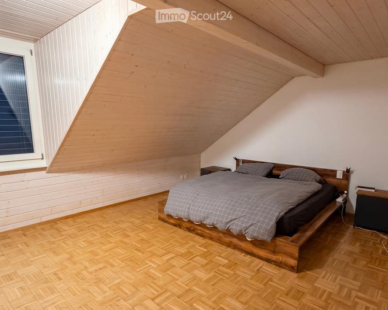 Cozy attic bedroom with wooden slanted ceilings, herringbone wooden flooring, and a large window. Features a wooden bed with a grey duvet and matching pillows, flanked by nightstands.