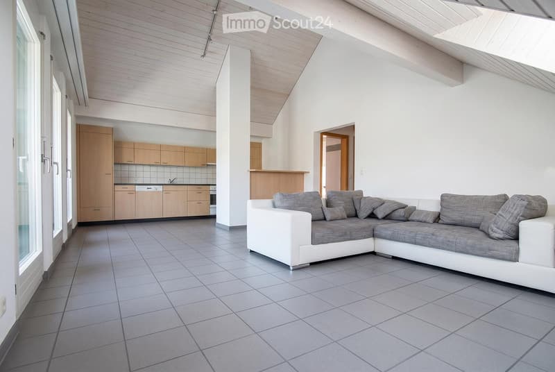 Modern living room with a white sectional sofa, grey cushions, light wooden cabinets, and a tiled floor. The room features large windows and a kitchen area with cabinets and appliances.