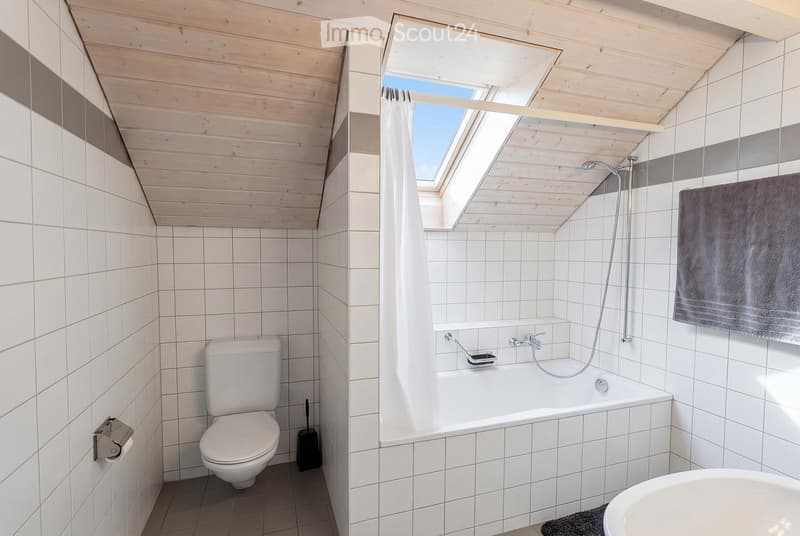 Modern bathroom with white tiles, wooden ceiling, skylight window, bathtub, toilet, and towel.
