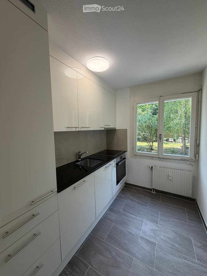 Modern kitchen with white cabinets, black countertop, sink, oven, and large windows overlooking a green area.