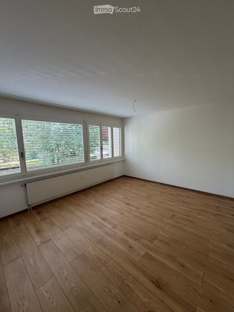 Empty room with wooden flooring, white walls, large windows with blinds, and a radiator under the windows.