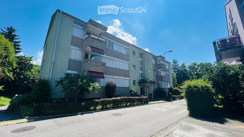 Multi-story residential building with balconies, surrounded by greenery and a clear blue sky.