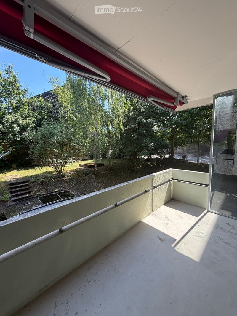 Balcony with a retractable awning, concrete floor, low walls, and a view of a green garden area with trees and a parked car.