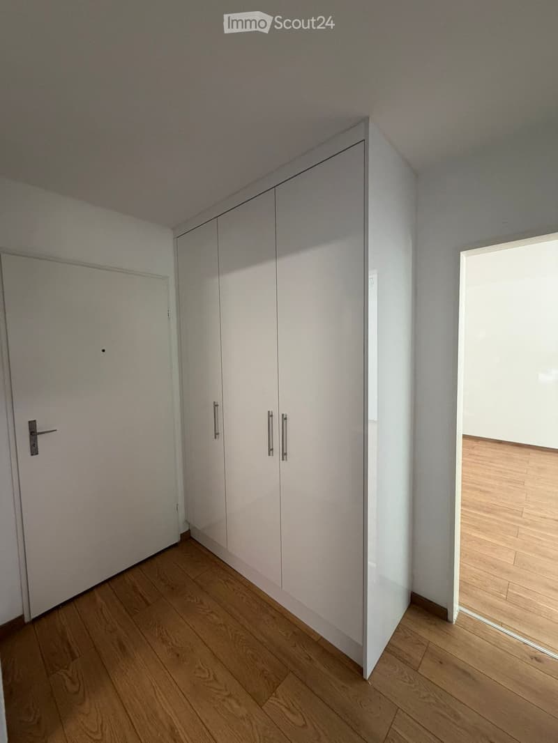 Empty room with light wooden flooring, white walls, and built-in white closet units with silver handles. A door is visible on the left side, and a window allows natural light to enter.