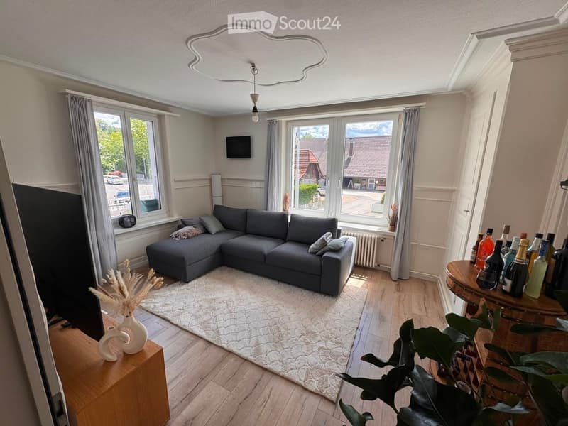 Modern living room with grey sectional sofa, coffee table, wooden floor, large windows, and decorative items.