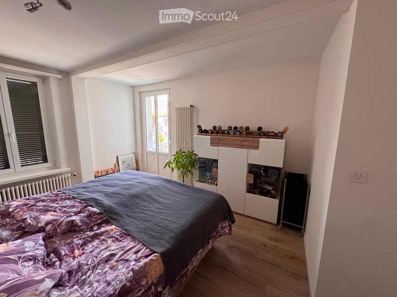 Bedroom with a double bed, a white wardrobe, a radiator, a picture frame, and a small plant. The room has wooden flooring and large windows with blinds.