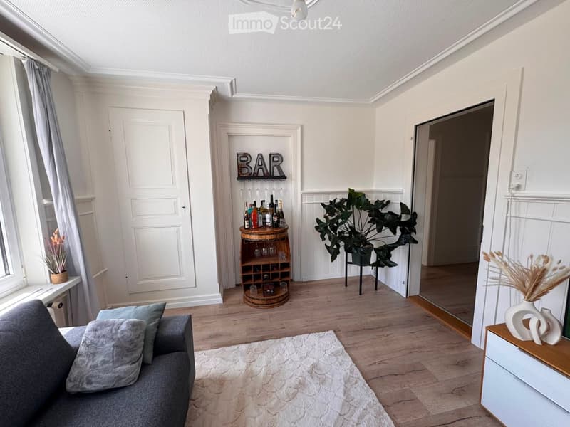 Living room with a grey sofa, wooden floor, white walls, a wine rack, a large plant, and decorative vases.