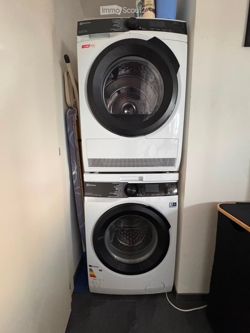 A compact bathroom with a stacked washing machine and dishwasher unit, white walls, and a dark floor.
