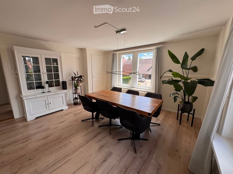 Bright dining room with wooden flooring, a large wooden dining table surrounded by black chairs, a white cabinet with glass doors, and potted plants near the windows.