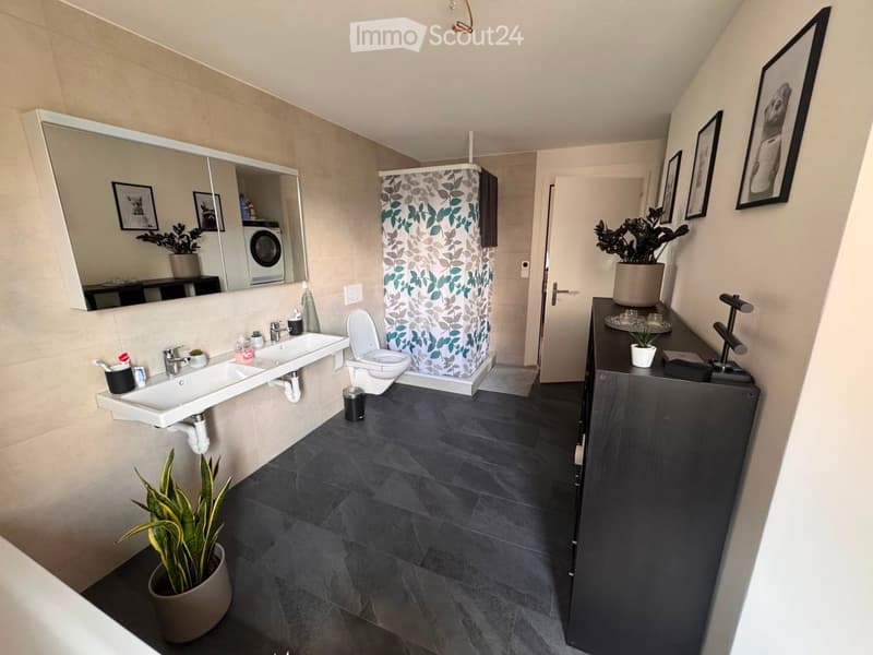 Modern bathroom with double sink, toilet, large mirror, dark tiled floor, and decorative plants.