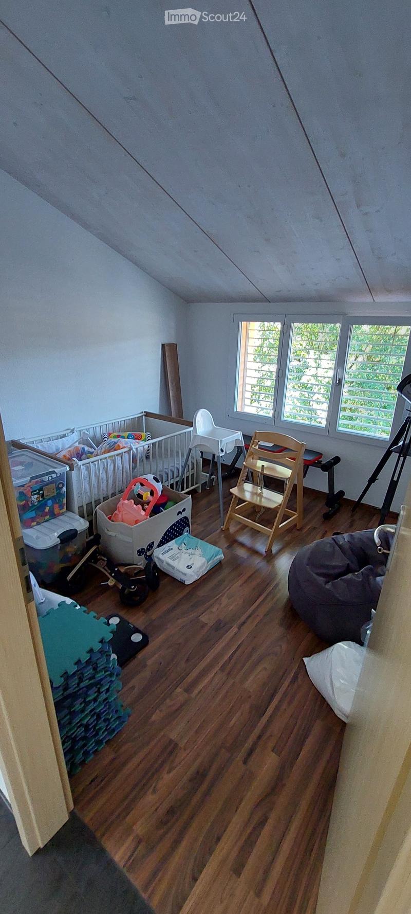 A well-lit attic room with wooden flooring, large windows, and various baby and toddler furniture and toys, including a crib, high chair, bean bag, and storage bins.