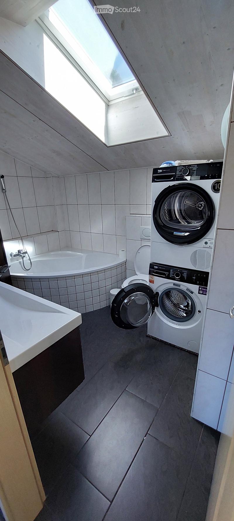 Modern bathroom with a bathtub, toilet, and washing machine. The room features white tiled walls and a skylight window at the top.