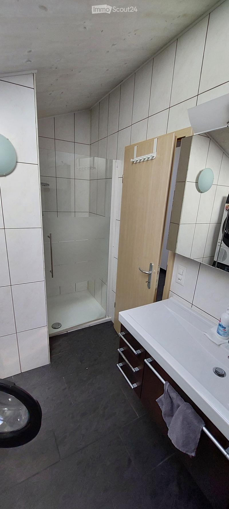Modern bathroom with white tiles, a glass-enclosed shower, a white sink with a wooden cabinet, and a mirror on the wall.