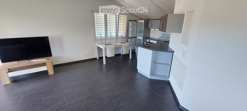 Open-plan living and kitchen area with dark wood flooring, white walls, and large windows with blinds. Features a television on a wooden stand, a small table with stools, a kitchen island, and modern kitchen cabinets.