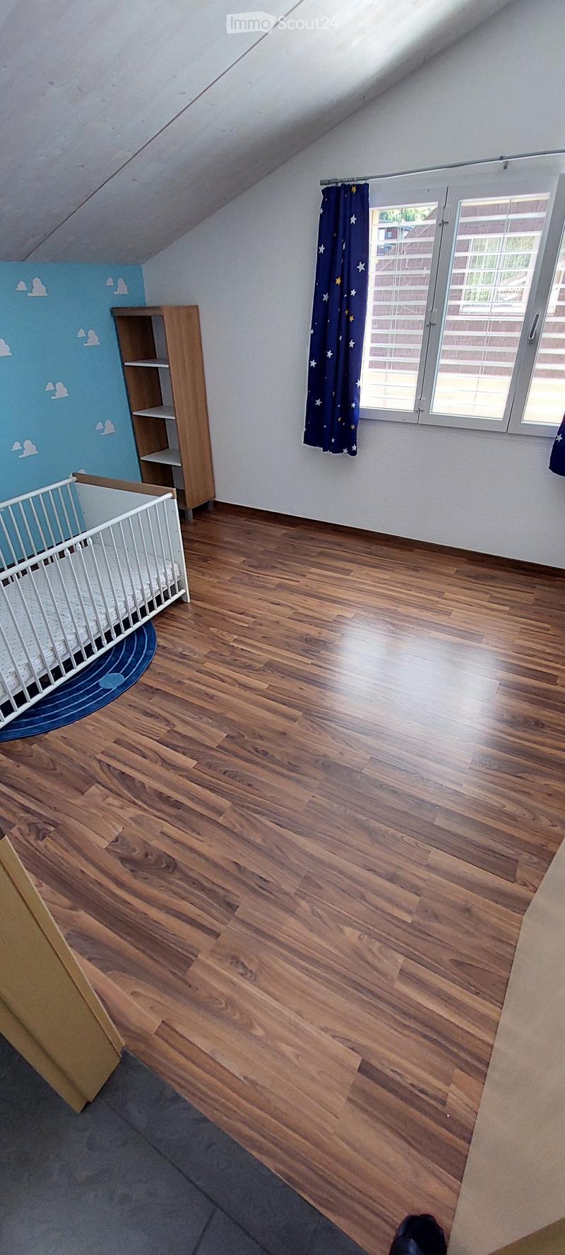 Bright, wooden-floored room with a crib, a wooden shelf, and a window with blue curtains featuring star patterns.