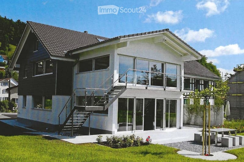 Modern two-story house with white exterior, large windows, and a raised ground floor accessed by stairs. Features a well-maintained lawn and small garden area with a bench and tree.