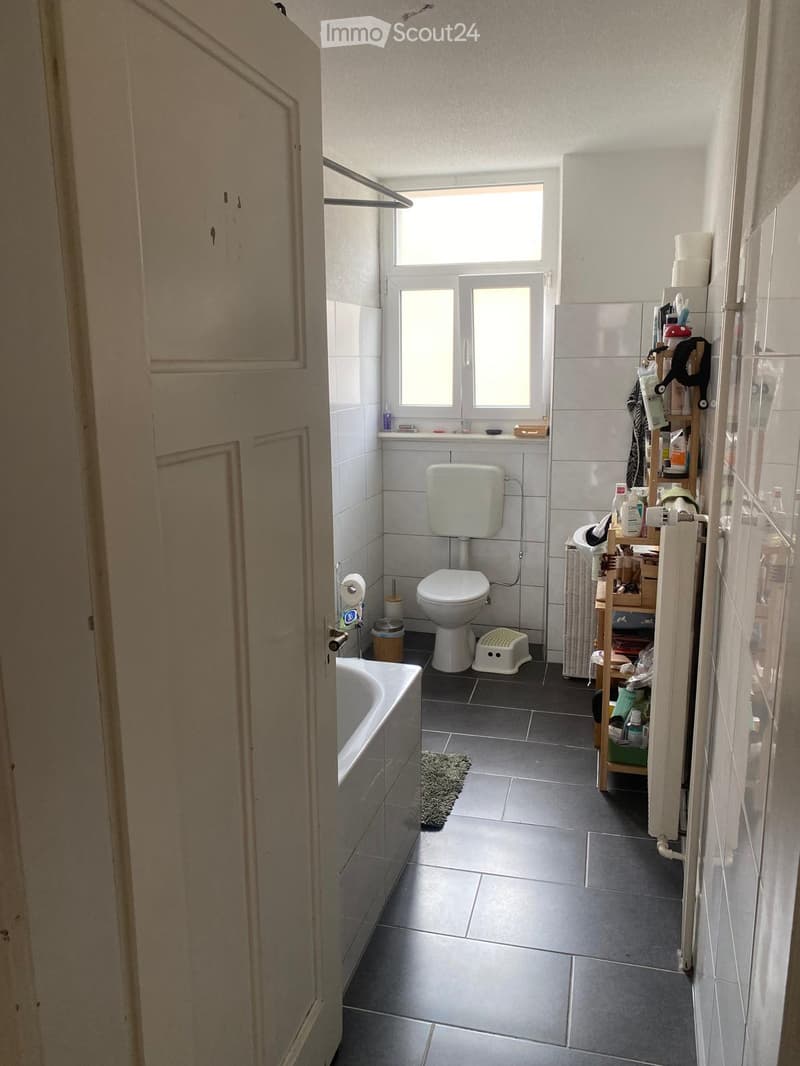Bathroom with a toilet, bathtub, sink, shower, shelf, stool, mirror, and toilet paper. The room has tiled walls and a dark tiled floor.