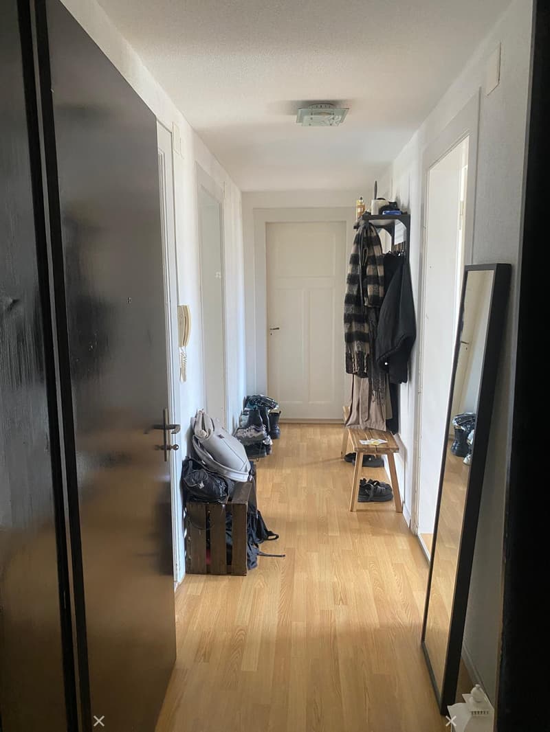 A narrow hallway with wooden flooring, white walls, and several doors. There are coats and shoes placed on small benches on either side.