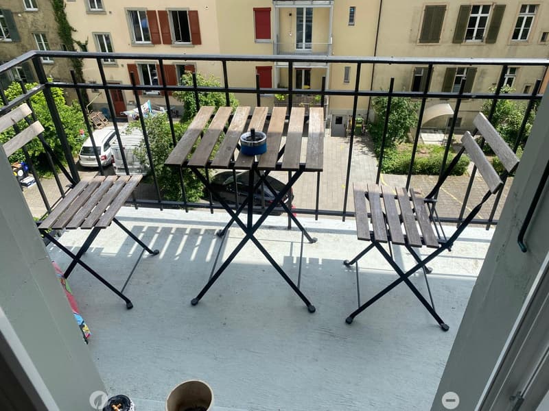 Balcony with a small wooden table and four folding chairs, overlooking a residential area with parked cars and greenery.