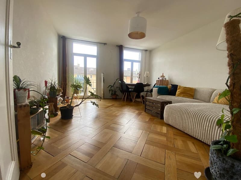 Modern living room with wooden flooring, large windows, a sectional sofa, a dining table with chairs, and several potted plants.