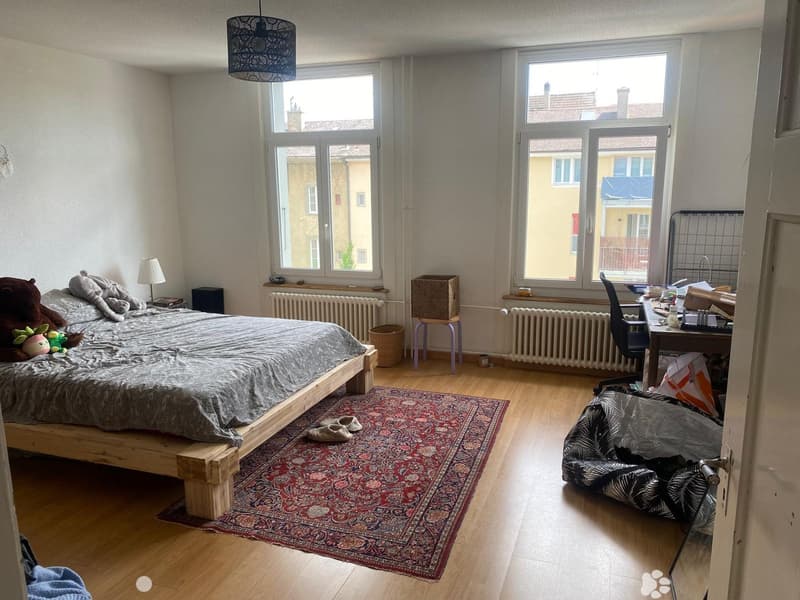 Cozy bedroom with wooden flooring, large windows, a bed with stuffed animals, a desk with a chair, a small lamp, a wicker basket, a colorful rug, and a beanie bag.