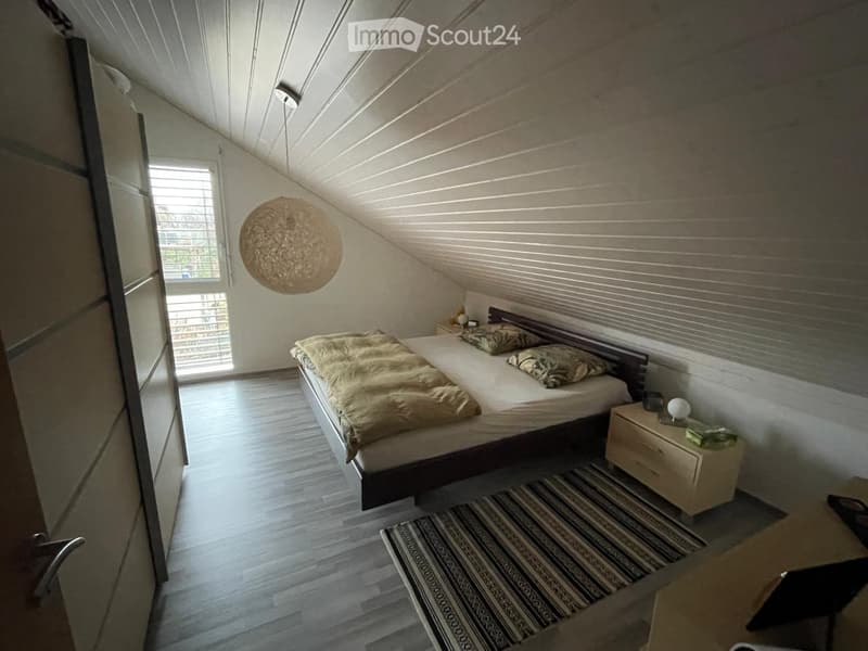 Cozy attic bedroom with wooden floors, slanted ceilings, and large window. Features a double bed with pillows and blankets, a nightstand with a lamp, and a patterned rug.