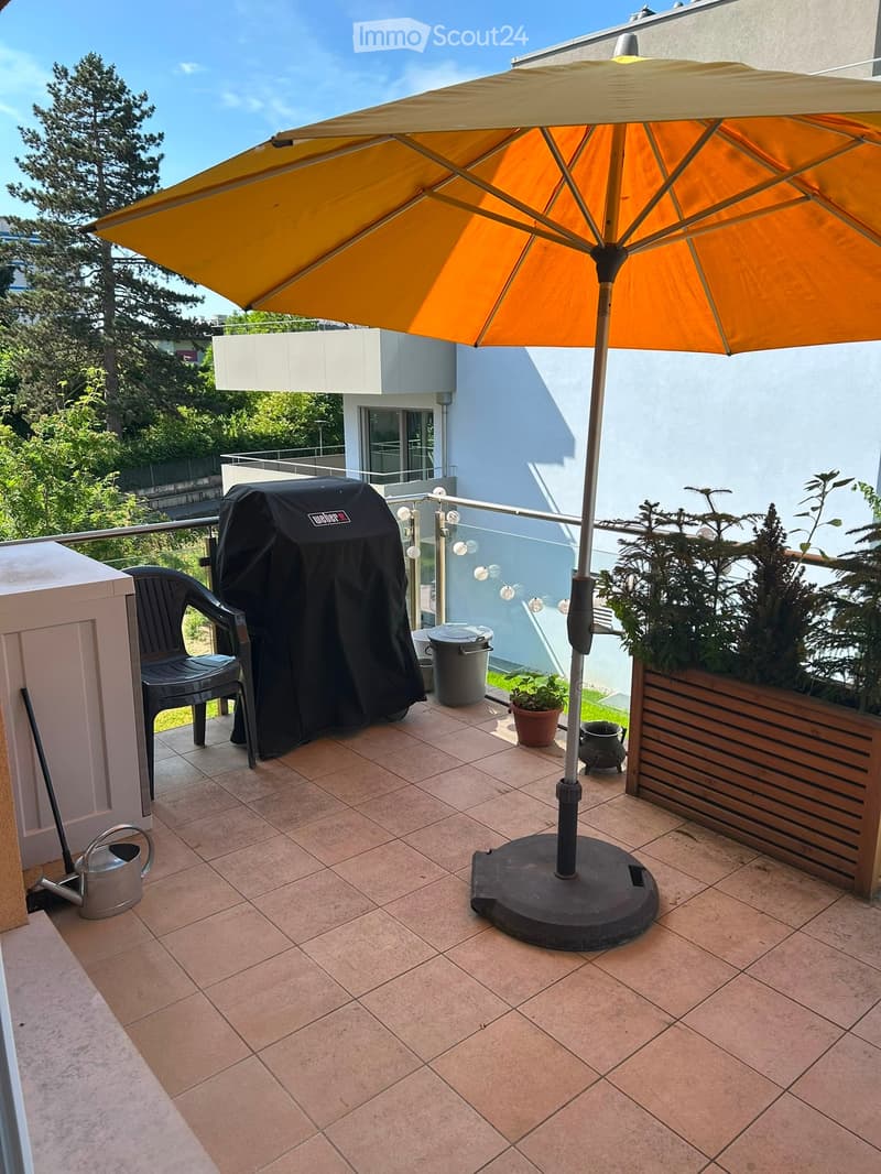 Balcony with tiled floor, large umbrella, black grill, chair, potted plants, and trash bin.