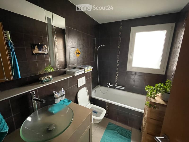 Modern bathroom with dark tiled walls, a glass sink, a bathtub, a toilet, a large mirror, and a window letting in natural light. There are towels, a small plant, and decorative items on the counter.