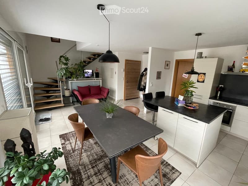 Open-plan living and dining area with a modern kitchen island, dining table with chairs, sofa, pendant lights, and plants.