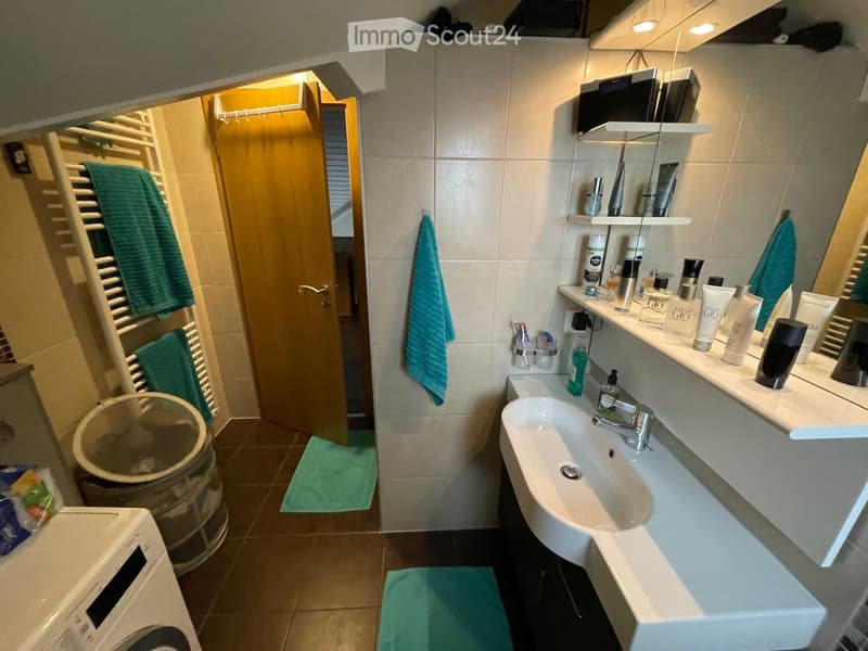 Modern bathroom with a white sink, mirror, shelves with toiletries, and a washing machine. Green towels are placed on the floor and a towel rack.