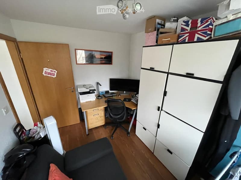 A furnished room with a desk, computer, printer, chair, sofa, and cabinet. The room has wooden flooring and white walls.